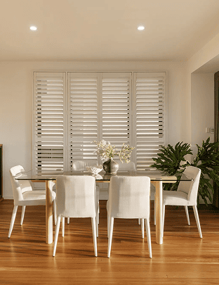 Modern dining room with white chairs and wooden floor, large window with shutters.